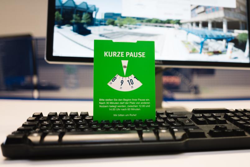 Eine grüne Parkscheibe, auf der "Kurze Pause" steht, steckt in einer Tastatur vor einem PC-Bildschirm.