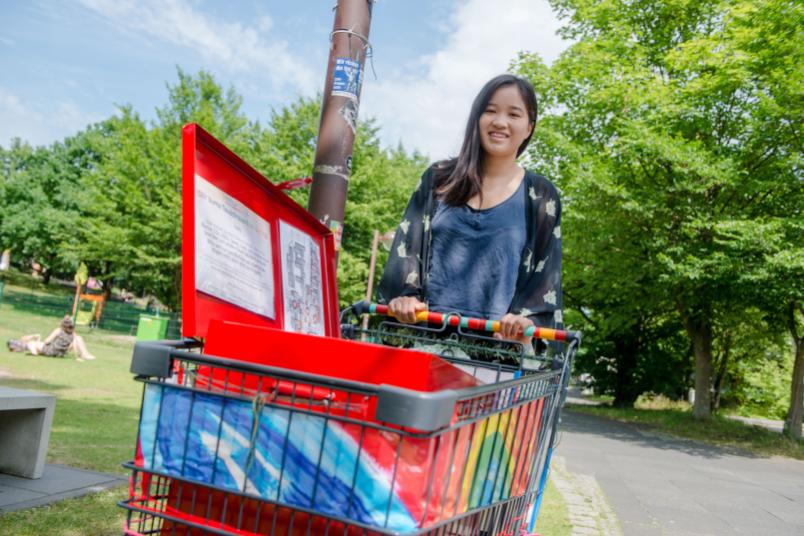 Eine Frau steht hinter einem bunten Einkaufswagen, in dem eine bunte Box steht.