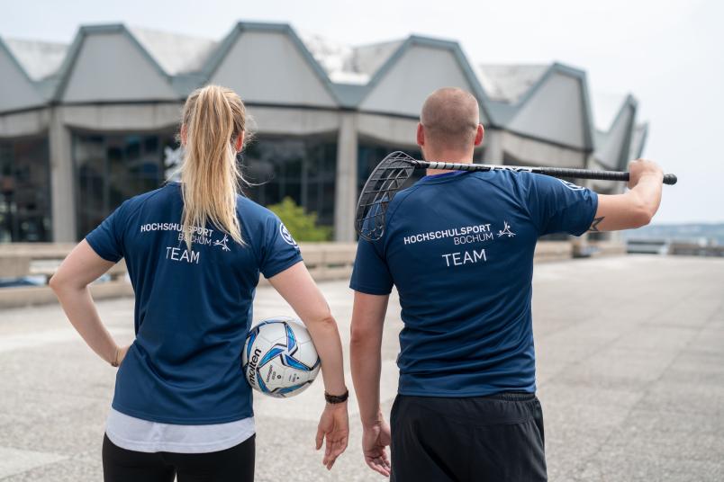Eine Frau mit Fußball unter dem Arm und ein Mann mit Hockeyschläger über der Schulter von hinten
