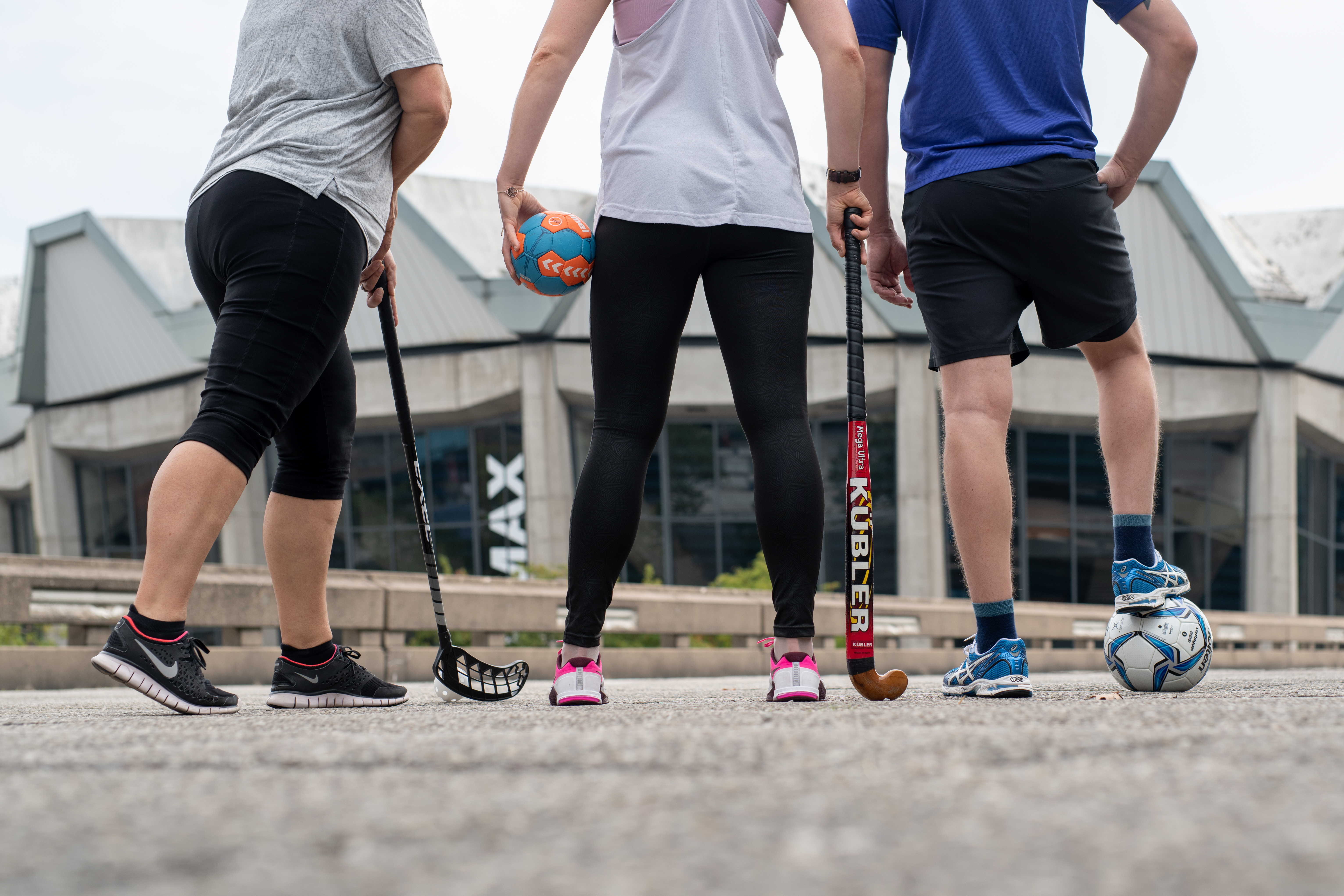 Drei Sportlerinnen und Sportler von hinten, mit diversen Sportgeräten