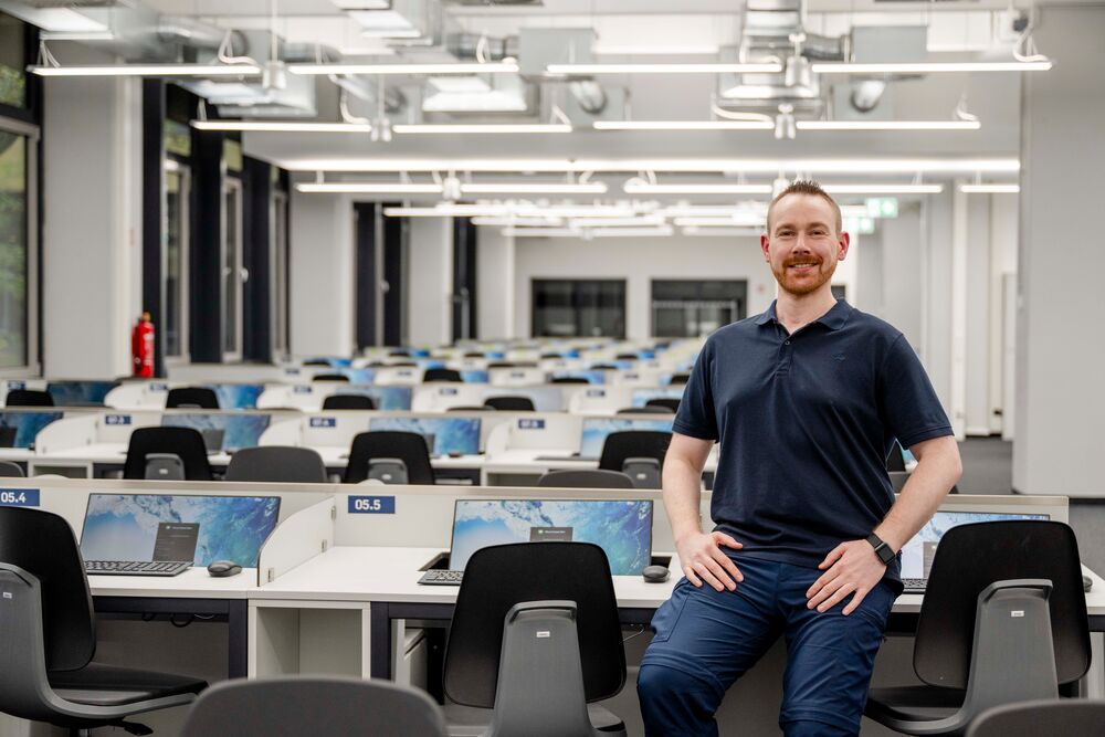 Mann sitzt auf einem Schreibtisch. Hinter ihm ein Raum mit Prüfungsplätzen.