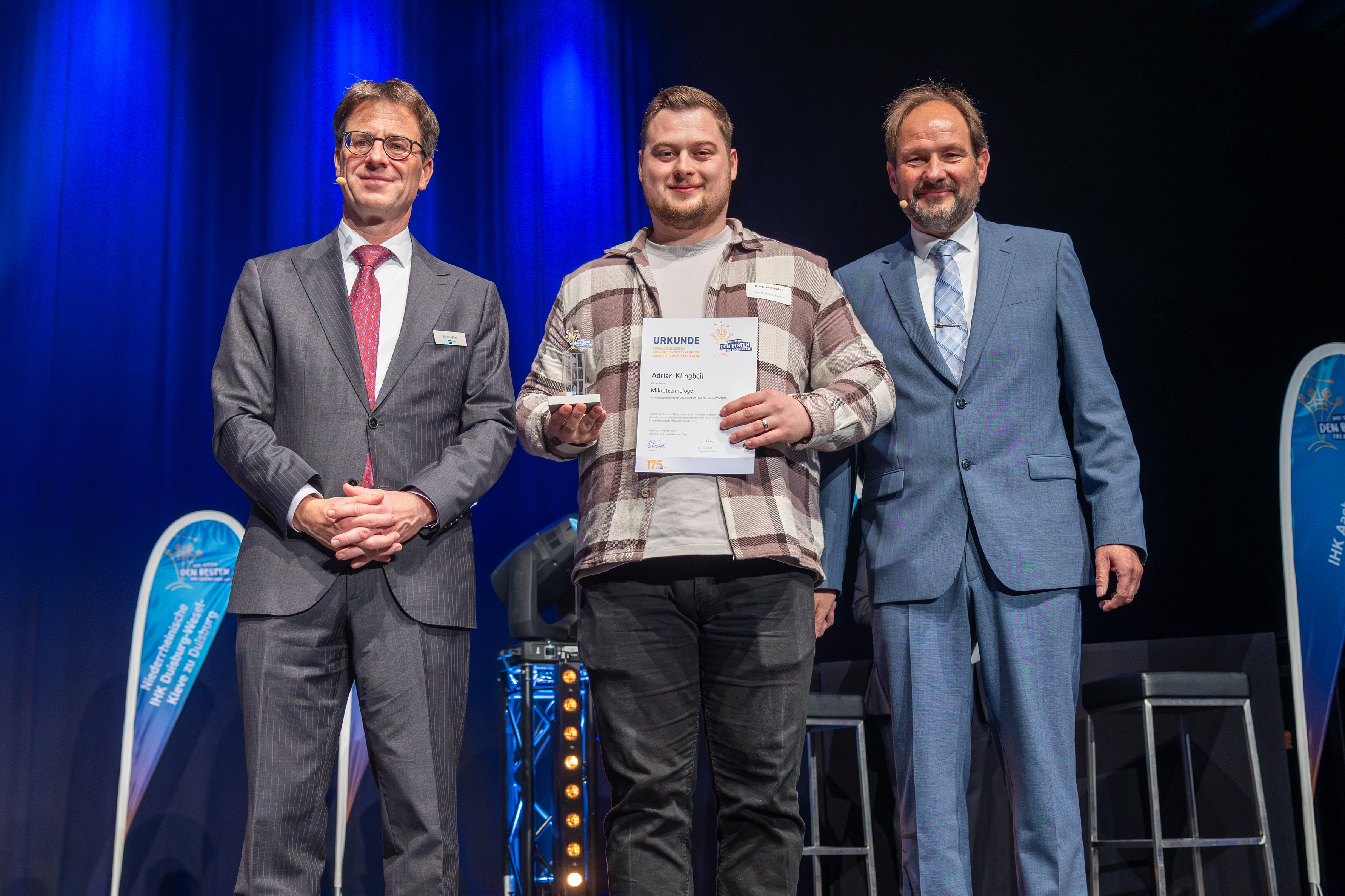 Drei Personen, einer davon mit Urkunde in der Hand