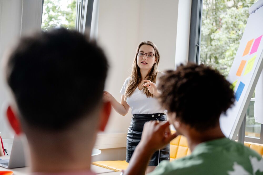 Lehrende steht an einem Flipchart und erklärt Studierenden etwas.