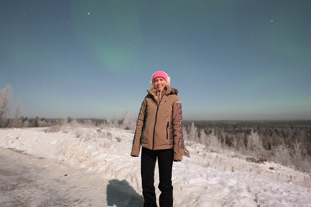 Frau in Wintersachen steht im Schnee. Hinter ihr sind Polarlichter zu sehen. 