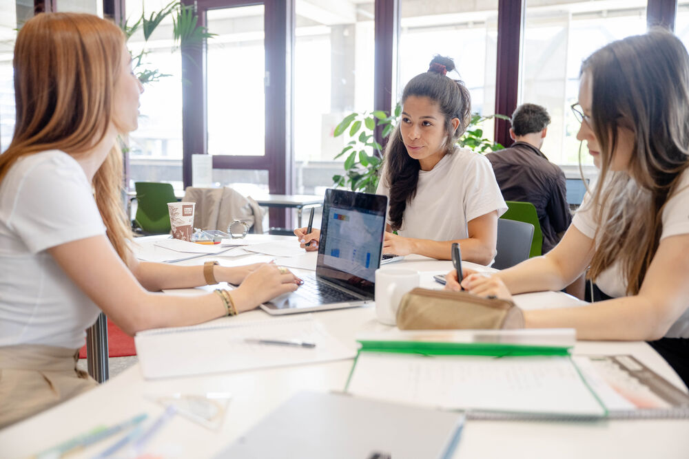 Drei Studentinnen lernen zusammen. Ein Laptop steht auf dem Tisch. Bücher liegen daneben. 