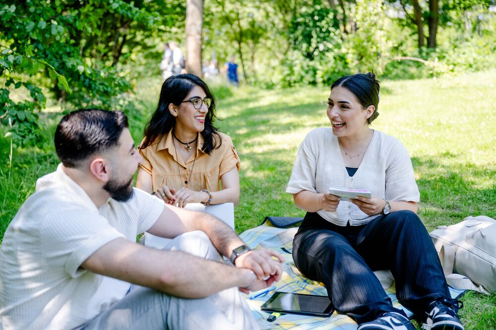 Studierende sitzen auf einer Wiese und unterhalten sich. Es scheint die Sonne.