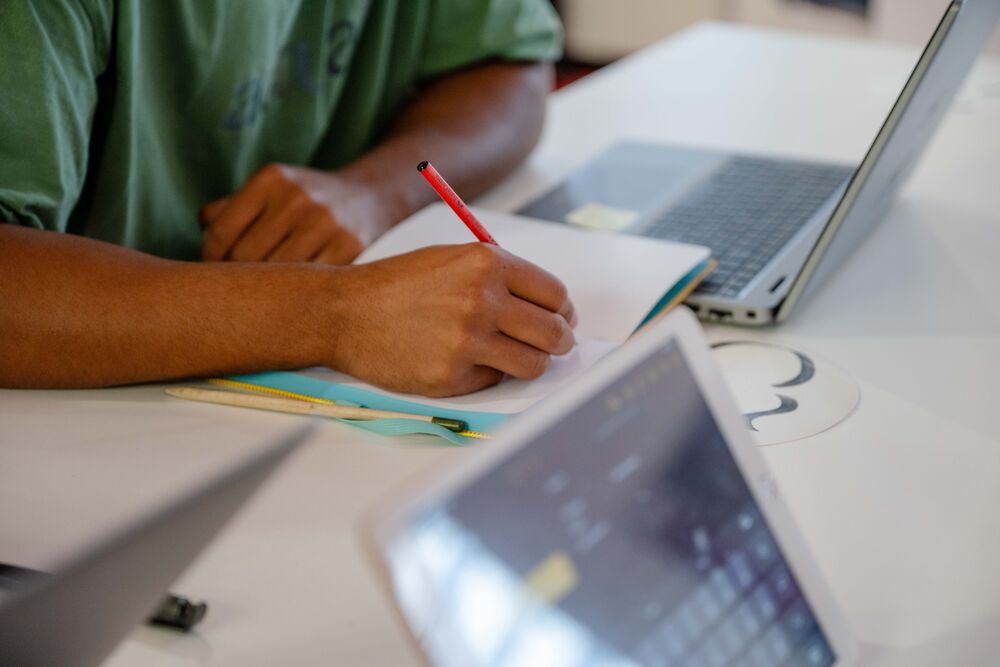 Student schreibt etwas auf. Es sind Laptops auf dem Tisch. 