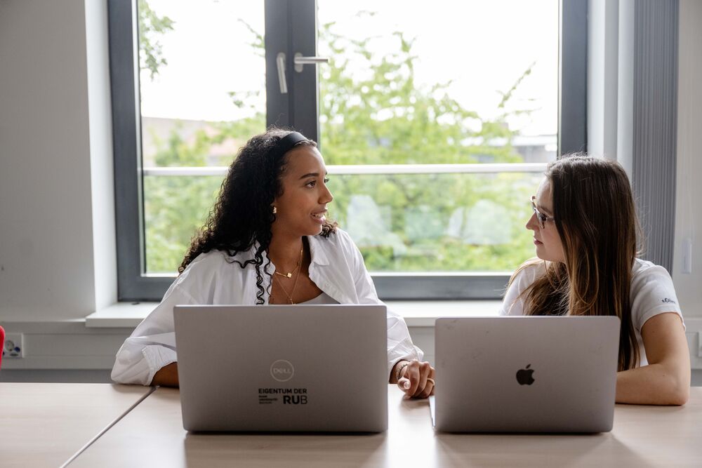 Zwei Frauen sitzt an zwei Laptops nebeneinander und unterhalten sich. 