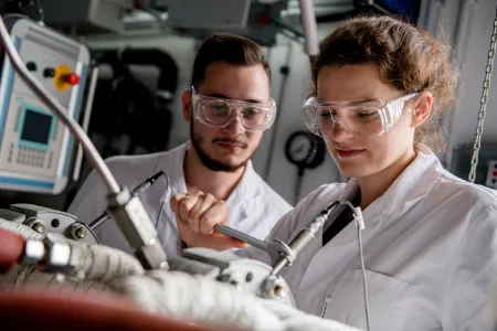 Ein Wissenschaftler und eine Wissenschaftlerin tragen Schutzbrillen und weiße Kittel und schauen auf eine Gerätschaft aus dem Maschinenbau.