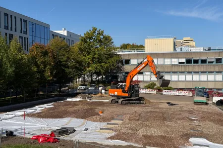 Ein orangener Bagger verteilt Schotter auf einer Fläche zwischen Gebäuden.