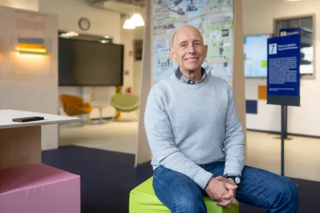 Mann im hellblauen Pulli auf einem hellgrünen Hocker vor einem Poster