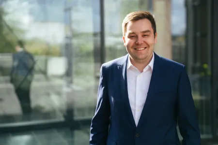 Ein junger, lachender Mann im weißen Hemd mit blauem Jackt vor einer spiegelnden Scheibe