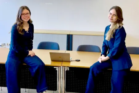 Zwei Studentinnen sitzen sich gegenüber auf Tischen. Beide sind in dunkelblau gekleidet.