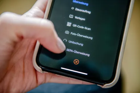 Nahaufnahme einer Hand, die ein Smartphone hält. Mit dem Daumen tippt sie auf den Menüpunkt "Überweisung".