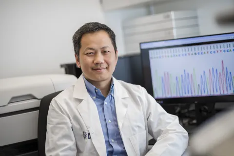 Ein Porträt eines jungen Mannes mit schwarzen kurzen Haaren und weißem Arztkittel. Im Hintergrund sieht man einen PC.