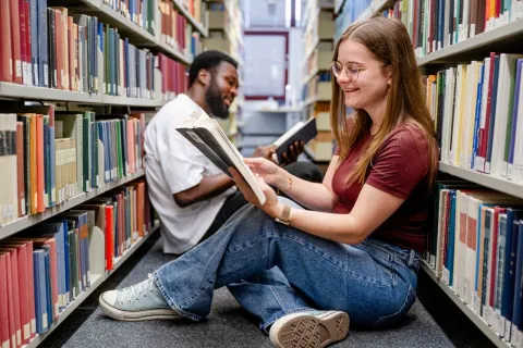 Zwei Studierende sitzen mit Büchern auf dem Boden der Bibliothek.