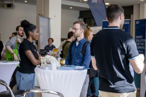 Ein Stehtisch an dem eine junge Frau und ein junger Mann stehen. Im Hintergrund sieht man Messetische und Aufsteller.