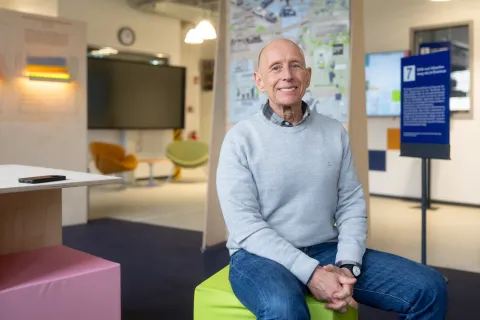 Mann im hellblauen Pulli auf einem hellgrünen Hocker vor einem Poster