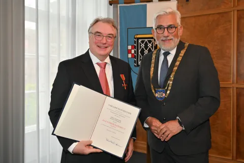 Zwei Männer im Anzug, einer zeigt eine Urkunde, der andere trägt eine Amtskette