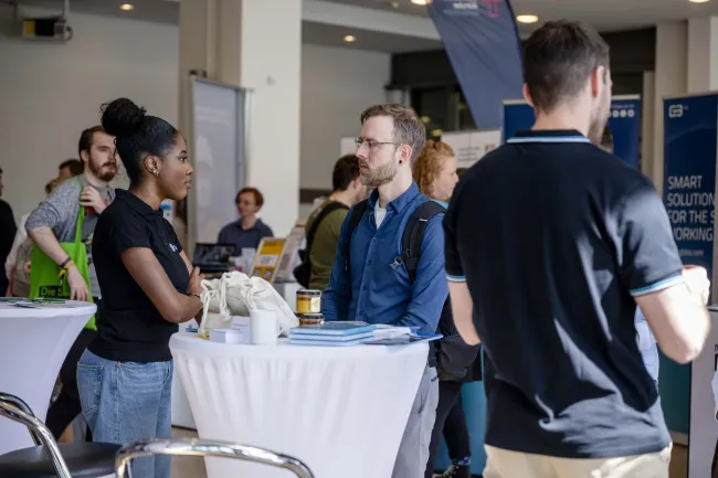 Ein Stehtisch an dem eine junge Frau und ein junger Mann stehen. Im Hintergrund sieht man Messetische und Aufsteller.