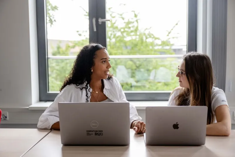 Zwei Frauen sitzt an zwei Laptops nebeneinander und unterhalten sich. 