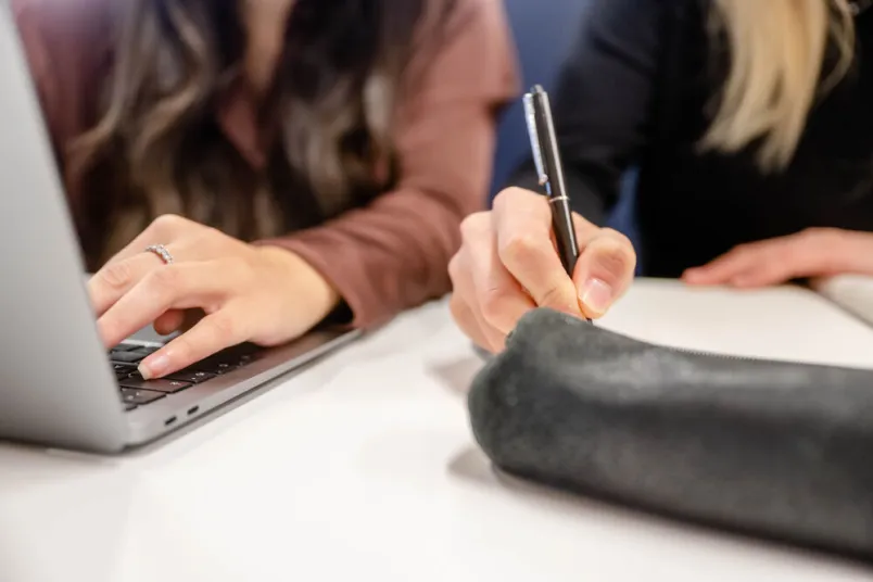 Hände zu sehen. Eine schreibt auf einer Laptoptastatur. Eine schreibt mit einem Stift auf Papier. 