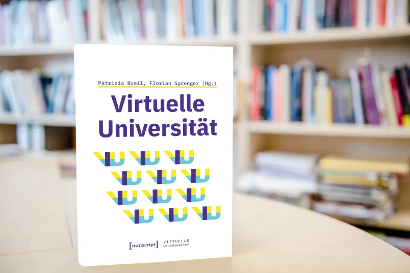 Buch mit Titel "Virtuelle Universität" steht auf Schreibtisch; dahinter im Hintergrund verschwommenes Buchregal