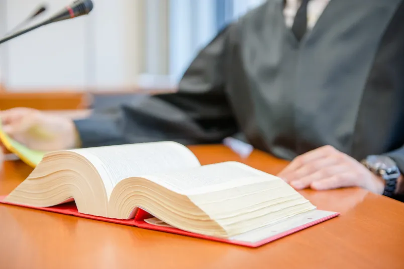 Ein Gesetzbuch liegt aufgeschlagen vor einer Person in Robe auf einem Tisch