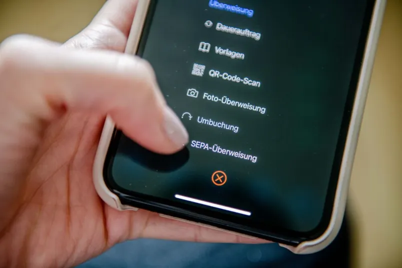 Nahaufnahme einer Hand, die ein Smartphone hält. Mit dem Daumen tippt sie auf den Menüpunkt "Überweisung".
