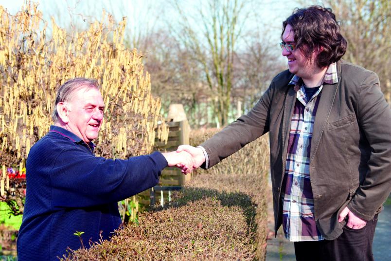 Zwei Männer reichen sich über eine Hecke hinweg die Hände.