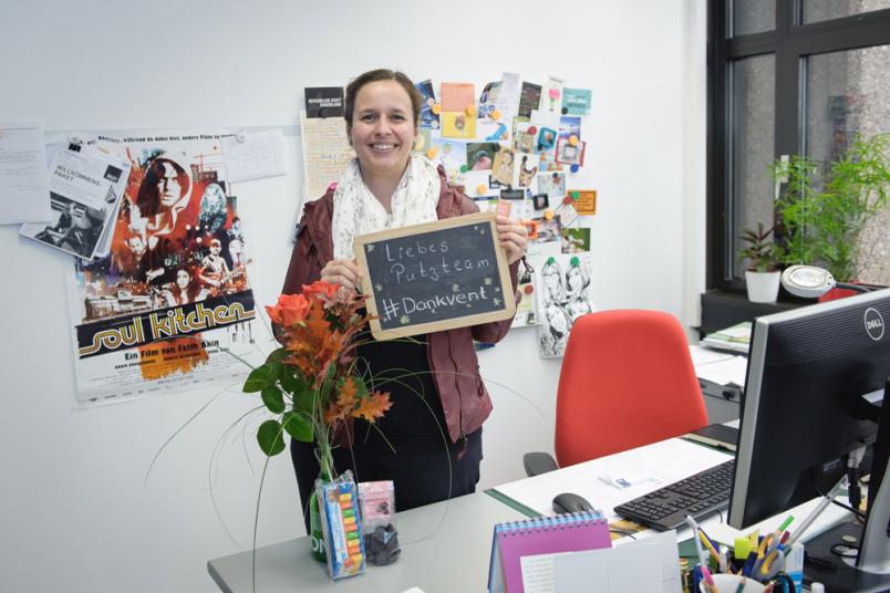 Eine Frau steht an ihrem Schreibtisch und hält eine Tafel vor sich, auf der steht "Liebes Putzteam #Dankvent".