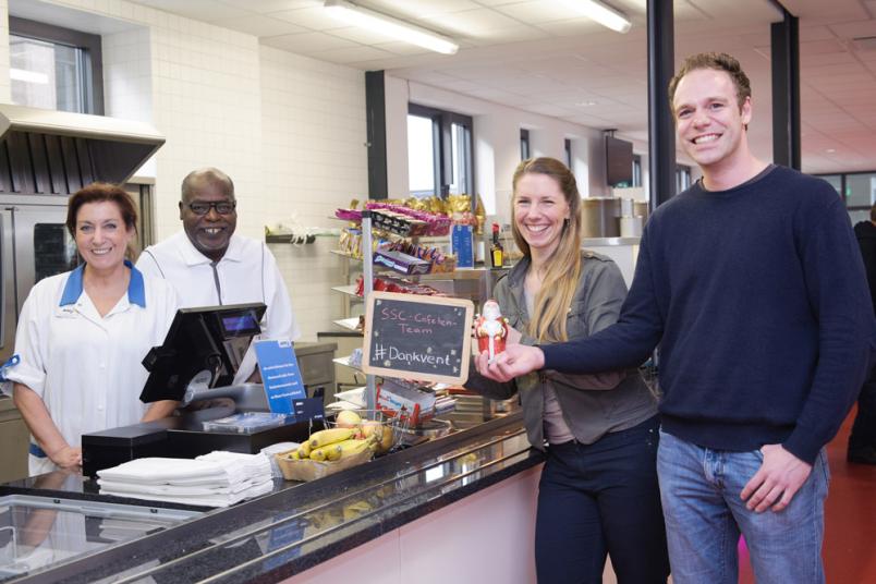 Ein Mann und eine Frau übergeben über die Theke ein Geschenk an zwei Mitarbeiter in der Cafeteria.