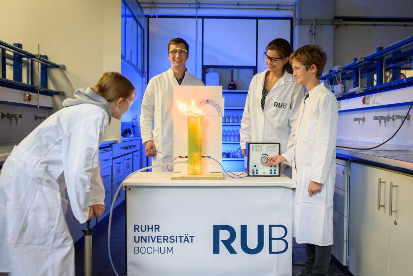 Zwei Kinder und zwei Forscher beobachten, wie eine Flamme aus einem Plexiglaszylinder schießt.