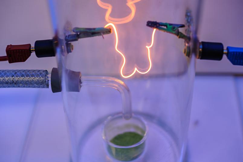 Ein glühender Draht in einem Plexiglasbehälter, darunter ein grünes Pulver