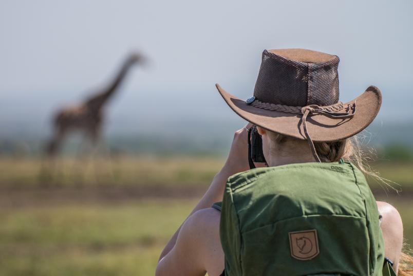 Eine Frau mit Rucksack und Hut von hinten beobachtet in der Ferne eine Giraffe.