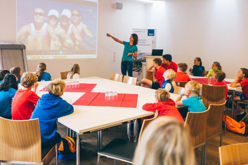 Eine Frau steht vorne in einem Raum und zeigt auf eine Beamerprojektion. Kinder sitzen an Tischen und hören zu.