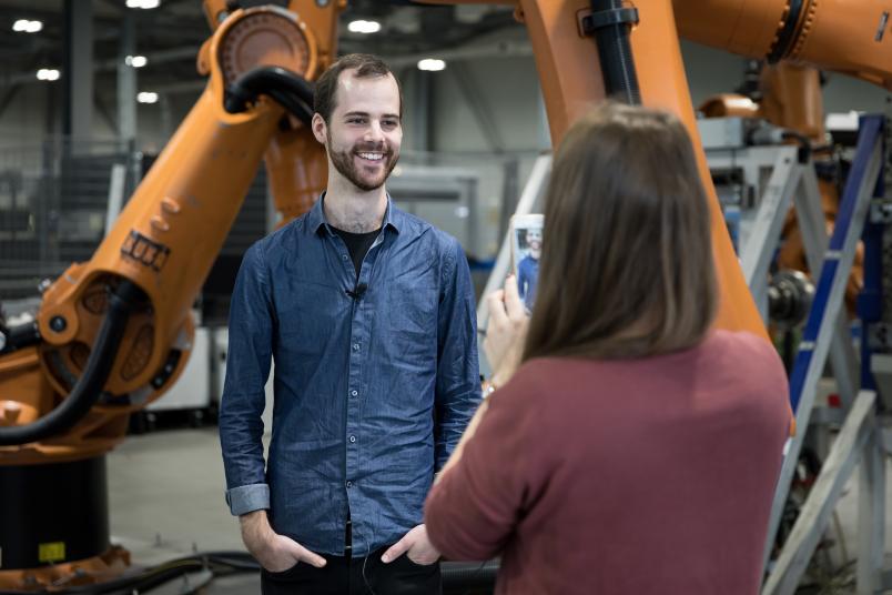 Ein Mann steht vor einem großen Roboterarm und lacht in eine Handykamera.