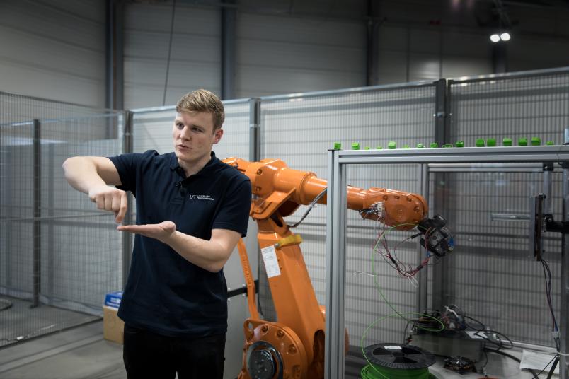 Ein Mann steht vor einem orangenen Roboter und gestikuliert erklärend.