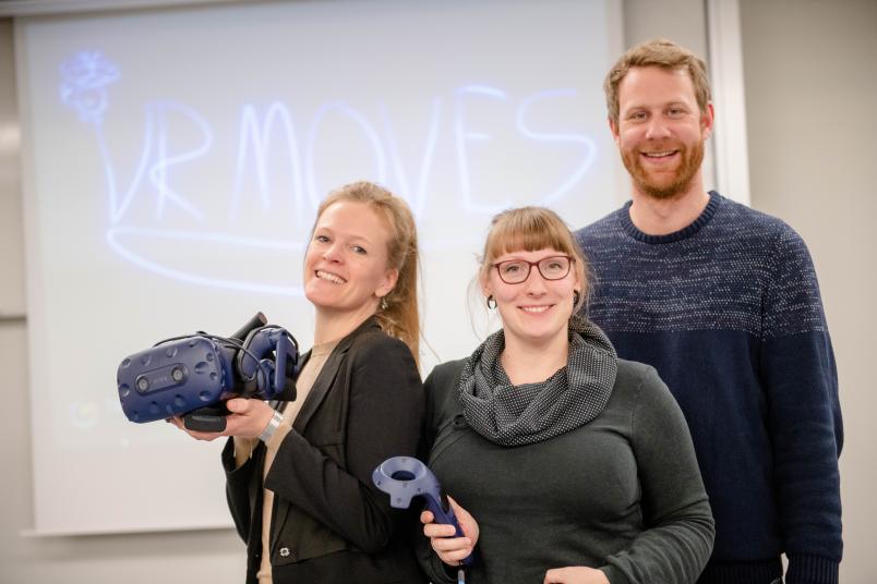Zwei Frauen und ein Mann, die Gegenstände in der Hand halten, die man benötigt, um virtuelle Realität zu erleben.