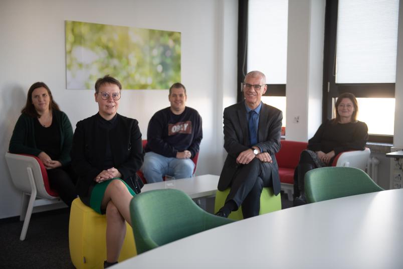 Fünf Personen sitzen um einen Couchtisch herum und schauen in die Kamera