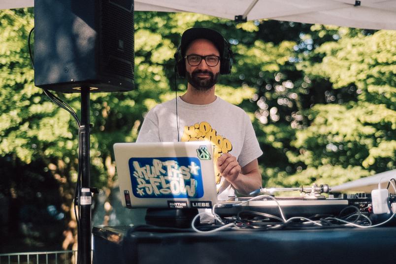 Ein Mann steht hinter DJ-Technik auf einer Bühne und schaut in die Kamera. Er trägt eine schwarze Cap und ein weißes Shirt mit gelber Schrift.