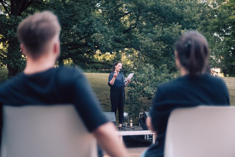 Eine Frau steht auf einer kleinen Bühne und spricht in ein Mikro. Sie trägt ein schwarzes T-Shirt. Fotografiert über die Schultern von zwei sitzenden Zuschauern.