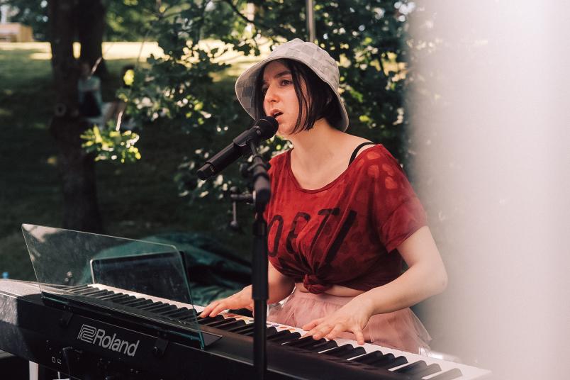 Eine Frau sitzt auf einer kleine Bühne an einem Keyboard und singt in ein Mikro. Sie trägt einen Hut und ein rotes T-Shirt.