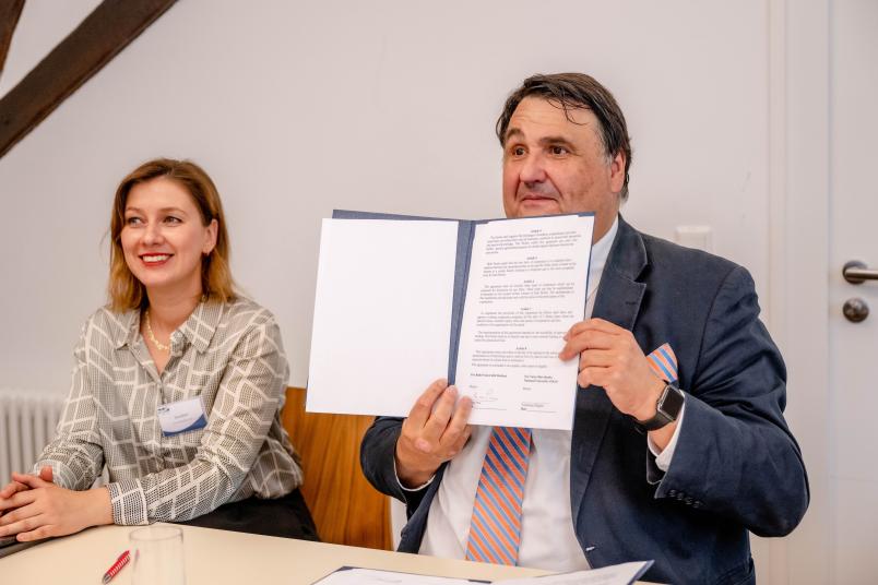 Stolz zeigt der Rektor den unterschriebenen Vertrag in die Kamera. Links neben ihm: Elena Resch, Organisatorin der Summer School