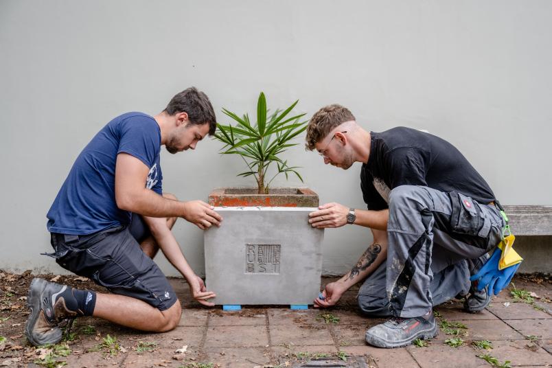 Zwei Personen arbeiten an einer Verkleidung für einen Betonkübel mit Palme