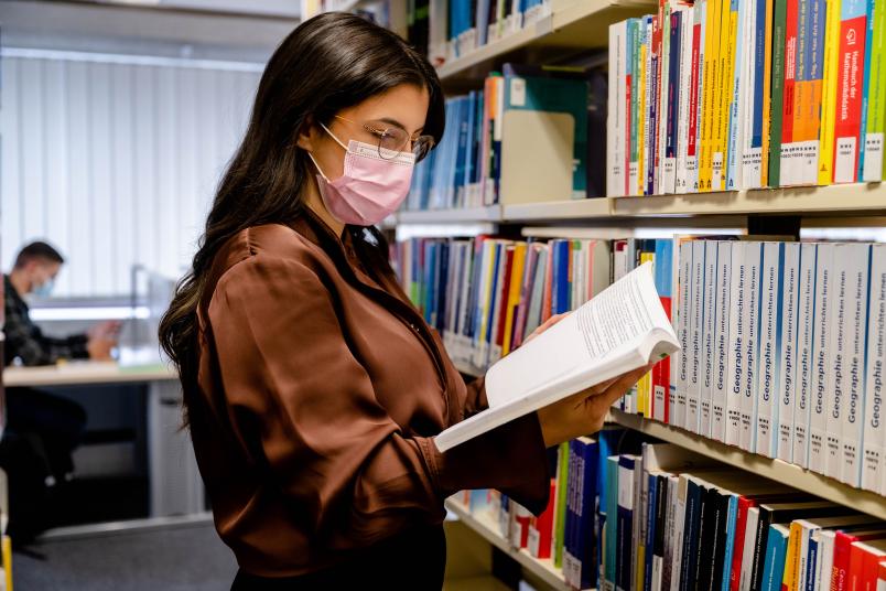 Studentin mit Mund-Nase-Schutz in der Bibliothek, im Hintergrund sitzt ein Studierender mit Maske an einem Tisch.