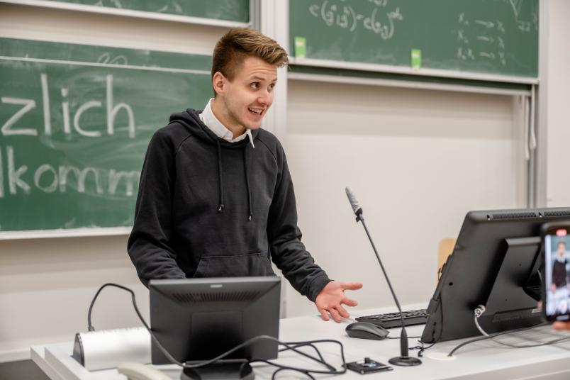 Max Lucks steht an einem Rednerpult in einem Hörsaal. Er spricht und gestikuliert.