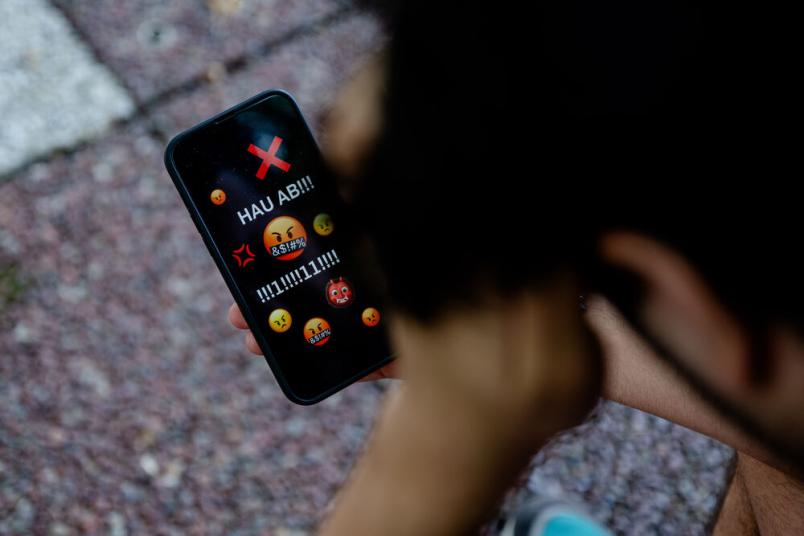 Detailaufnahme von einer Person, die ein Smartphone in der Hand hält. Darauf zu sehen sind wütende und schimpfende Emoticons und die Phrase "HAU AB!!!"