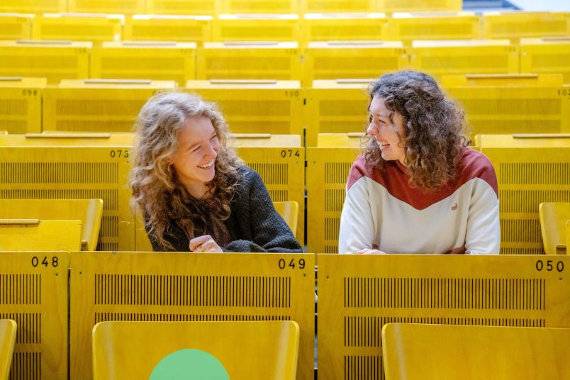 Charlotte Auel und Clara Bückert sitzen in einem Hörsaal und lachen sich an.