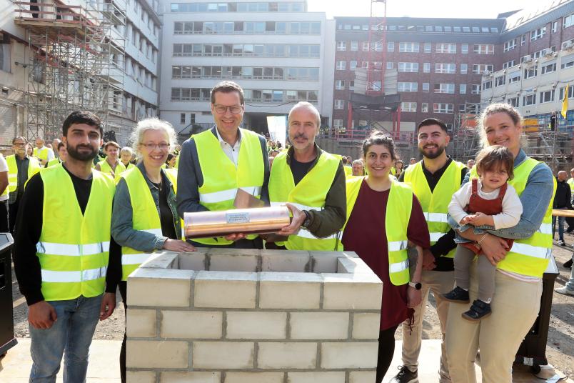 Mehrere Personen in Warnwesten auf einer Baustelle, zwei davon halten einen Metallzylinder in den Händen